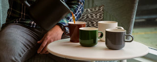 Acme mugs on a table with coffee being poured into them from a coffee carafe